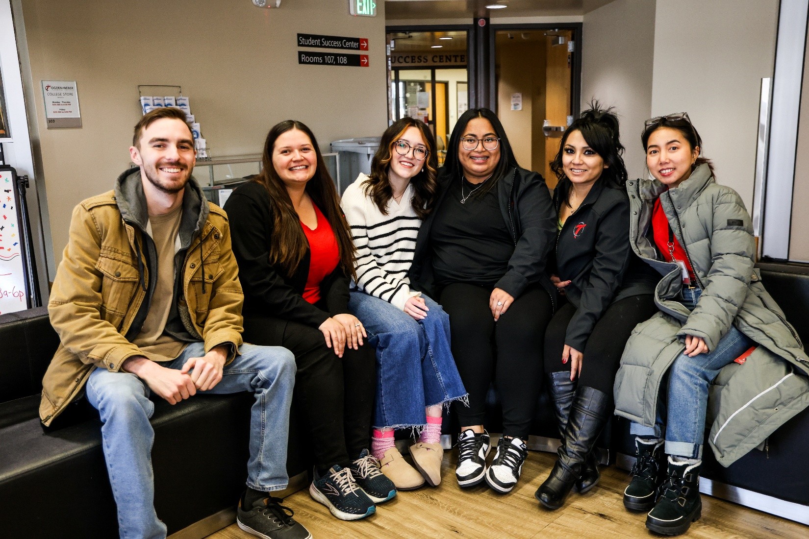 A group of six people sitting on black lounge seating posing for a picture inside a campus building, wearing winter clothing and casual attire, with signage for a Student Success Center visible in the background.